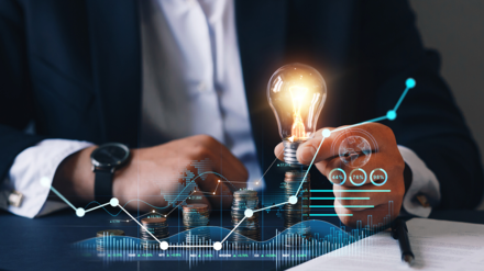 Mann im Anzug sitzt an einem Tisch und hält eine Glühbirne. Im Vordergrund wird eine Grafik gezeigt, welche den Anstieg der Energiepreise veranschaulichen soll