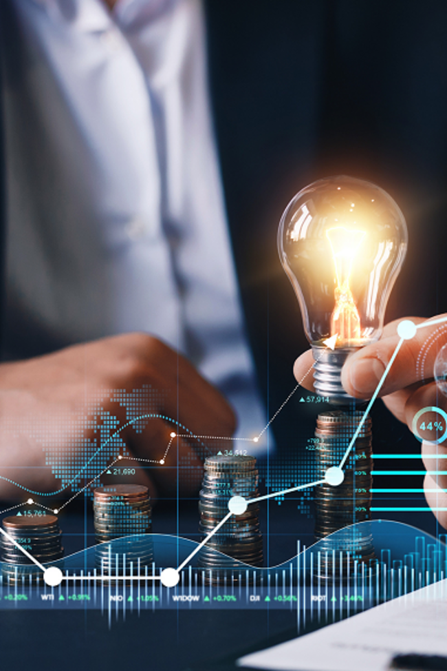 Mann im Anzug sitzt an einem Tisch und hält eine Glühbirne. Im Vordergrund wird eine Grafik gezeigt, welche den Anstieg der Energiepreise veranschaulichen soll