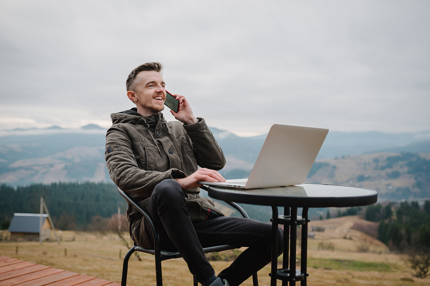 Mann sitzt am freien an kleinem Tisch und telefoniert, mit Laptop vor sich